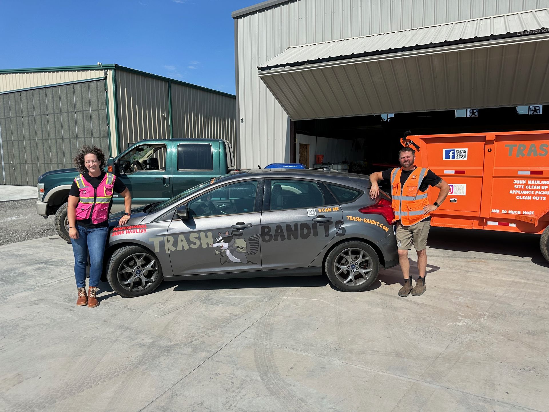 Israel and Jennifer beside the gray Trash Bandits car, with the orange trailer and open airplane hangar behind them